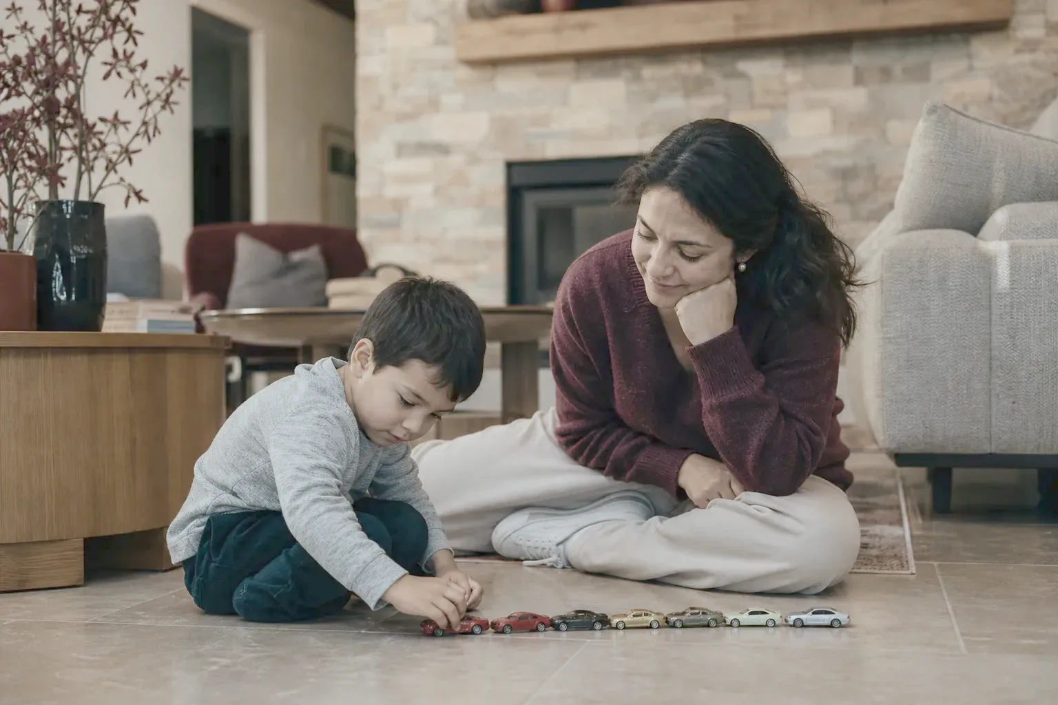 A mother sitting on the floor watching her son line up toy cars